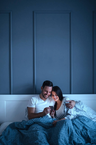 Young couple cuddling in bed while browsing the internet on their phone. - Foto, afbeelding