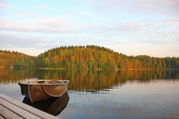 the boat is tied next to a wooden pier, smooth clear water of the lake, the shore covered with forest, autumn, Karelian nature, reflection in the water - Foto, Imagem