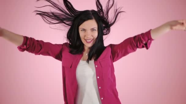Energy girl on pink background in studio. She wears colorful shirt. Black hair in tail is flying from moving. Slow motion - Video