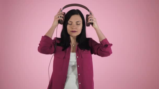 Beautiful young brunette woman in headphones listening to music and singing on color background. Happy girl in pink shirt. - Metraje, vídeo