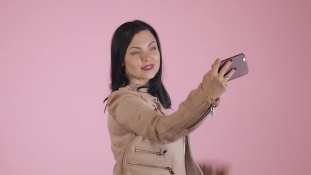 Smiling happy brunette woman in pink jacket making selfie on smartphone over colorful background. - Filmati, video