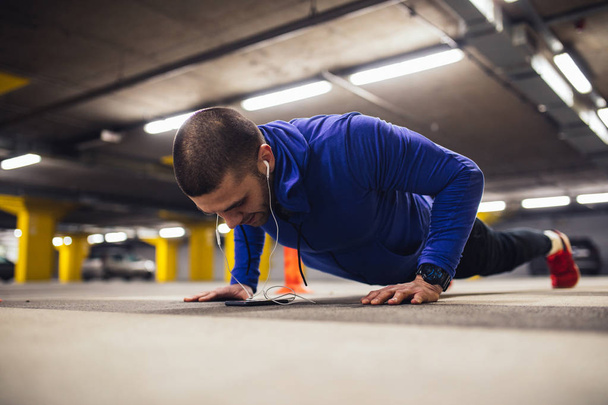 A young man doing pushups inside an urban garage while listening to music on his phone. - Foto, immagini