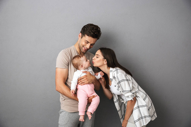 Portrait of a happy young family with their little baby girl isolated over gray background, kissing - Photo, image
