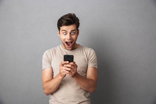 Happy cheerful guy holding smartphone and shouting with excitement isolated over gray background - Φωτογραφία, εικόνα