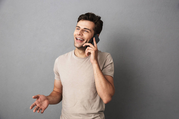 Portrait of a happy young man in t-shirt isolated over gray backgound, talking on mobile phone - Foto, imagen