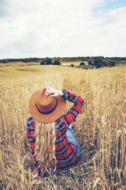 Zadní pohled na blond žena nosí klobouk sedí v žlutém poli pšenice  - Fotografie, Obrázek