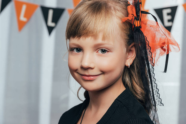 Porträt eines entzückenden lächelnden Kindes, das in die Kamera blickt und zu Hause Halloween-Fahnen aufhängt - Foto, Bild