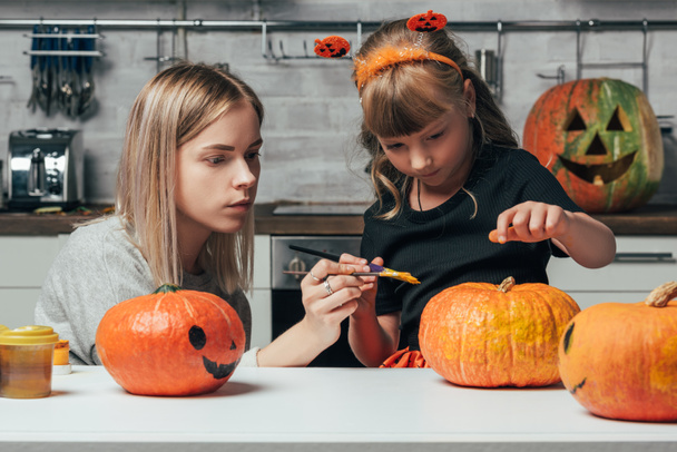young woman and little sister painting pumpkins for halloween together in kitchen at home - 写真・画像