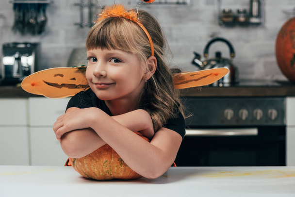 little kid in halloween costume standing at tabletop with pumpkin in kitchen at home - Photo, Image