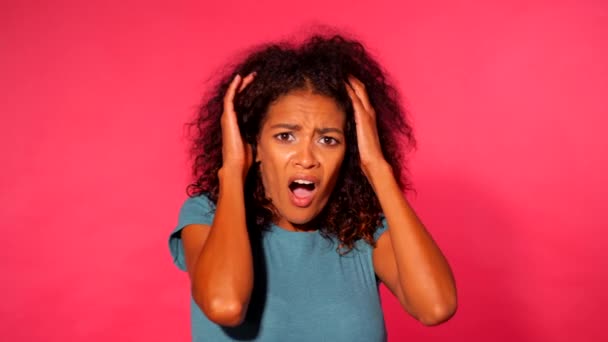 Frightened african american woman with curly hair in green t-shirt afraid of something and looks into the camera with big eyes full of horror over pink wall background. - Materiaali, video