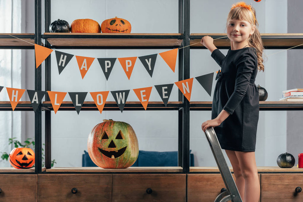 Niedliches Kind auf Leiter hängt Fahnen mit fröhlicher Halloween-Inschrift zu Hause - Foto, Bild
