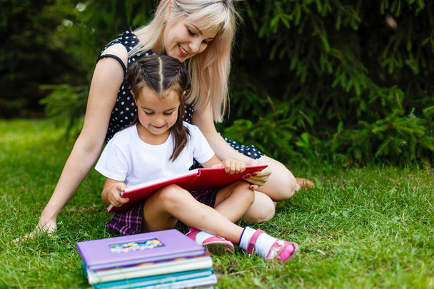 Mooie jonge moeder leesboek voor haar dochtertje zittend op de groene weide - Foto, afbeelding