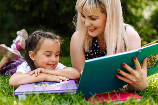 Bonita jovem mãe lendo livro para sua filha deitada no prado verde
 - Foto, Imagem