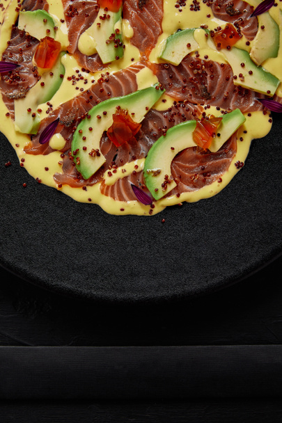 top view of delicious sliced fish and avocado in sauce on black plate - Foto, Bild