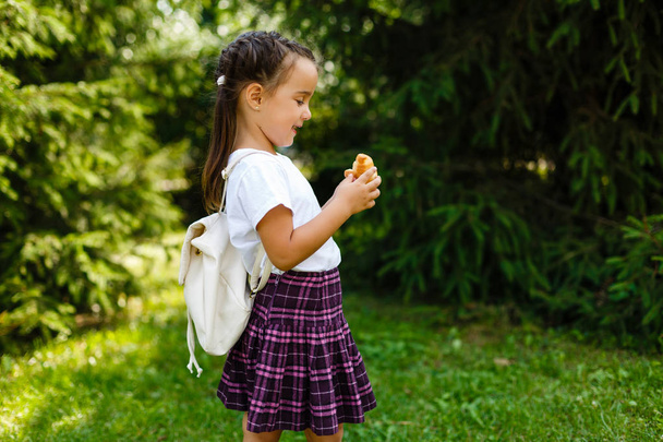 Una colegiala comiendo croissants en el parque. Volver a la escuela al aire libre
 - Foto, Imagen