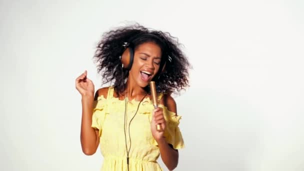 Young cute african american woman in yellow dress singing and dancing with hair dryer instead microphone at white background. Modern trendy girl with afro hairstyle. - Séquence, vidéo