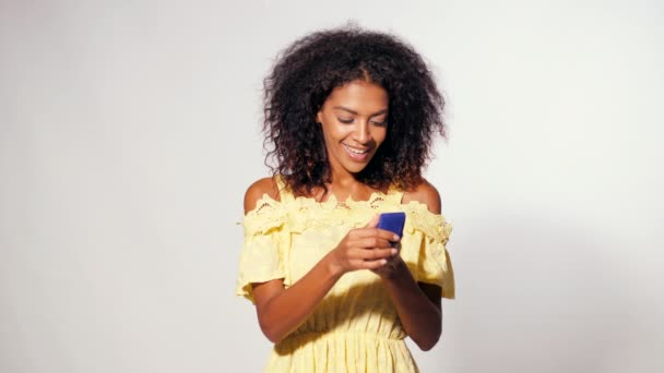 Pretty african-american young woman in yellow dress holding small jewelry box on white wall background. Girl smiling, she is happy to get present, offer of marriage. - Imágenes, Vídeo
