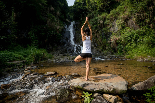 Young woman doing yoga near waterfall in forest - Фото, зображення