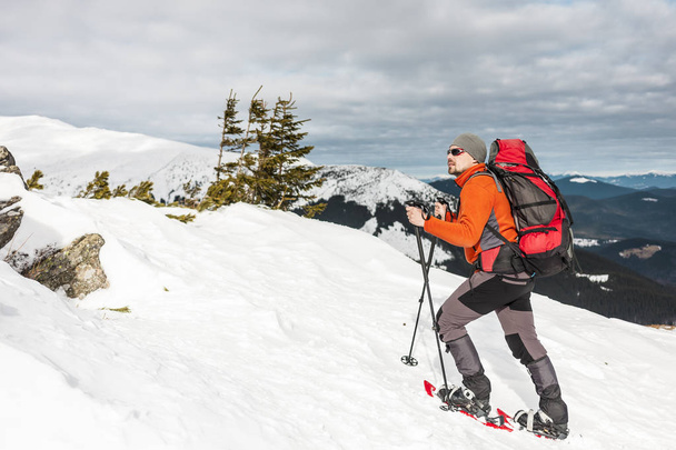 Winter climbing the mountain. A man in snowshoes is climbing to the top. Winter ascent. A mountaineer with a backpack and trekking sticks. Equipment for winter hiking. - Photo, Image