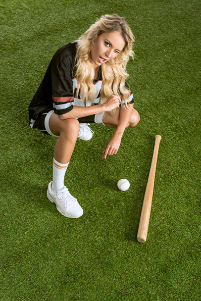 vista de ángulo alto de la joven atlética en uniforme de fútbol americano con bate de béisbol sentado en la hierba y mirando a la cámara
 - Foto, Imagen