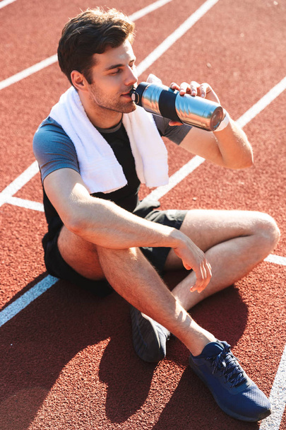 Esportista exausto terminou de correr no estádio, descansando, bebendo de uma garrafa de água
 - Foto, Imagem