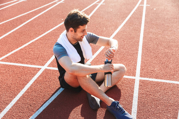 Le sportif épuisé a fini de courir au stade, se reposant, tenant la bouteille d'eau, regardant smartwatch
 - Photo, image