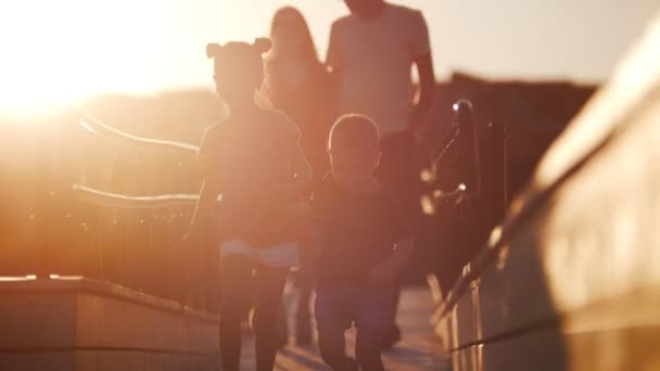 Młode rodziny pieszo na moście przed piękny zachód słońca. Dzieci działać dalej. Rodzice za robótką, trzymając się za ręce. - Materiał filmowy, wideo