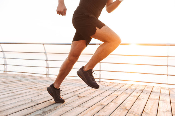 Piernas primer plano de hombre deportivo muscular 20s en chándal corriendo a lo largo del muelle o paseo marítimo en la playa por la mañana
 - Foto, Imagen