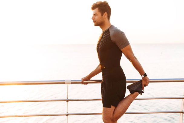 Image of caucasian man 20s in tracksuit working out and stretching legs on pier or boardwalk at seaside in morning - Φωτογραφία, εικόνα
