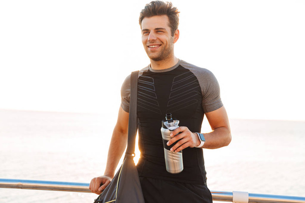 Photo of happy young man 20s in tracksuit with sporty bag walking along pier or boardwalk at seaside in morning and holding metal thermos mug - Fotografie, Obrázek