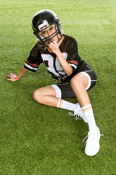angry young woman in american football uniform sitting on grass and looking at camera - Foto, afbeelding