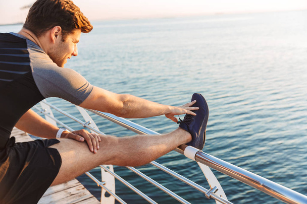 Image of young sporty man 20s in shorts and t-shirt raising leg on railing, and stretching body on boardwalk at seaside in morning - Фото, зображення