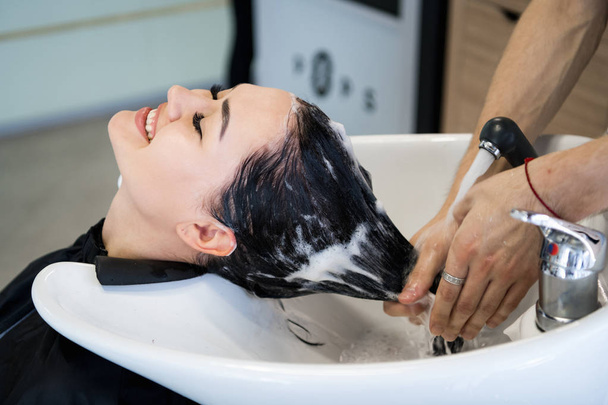 Процедура ухода за волосами в салоне красоты. Парикмахер расчесывает волосы женщин, распространяя лечебную маску или кондиционер
 - Фото, изображение