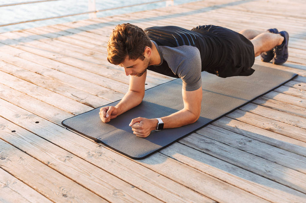 Photo of concentrated sportsman 20s in tracksuit lying on mat and doing plank while working out on wooden pier at seaside in morning - Photo, Image