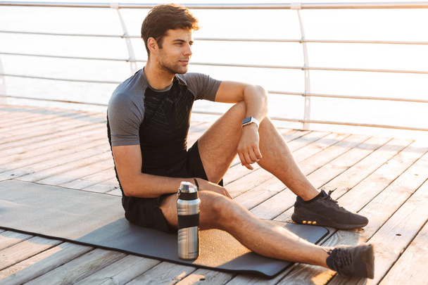 Photo of adult sportsman 20s in tracksuit sitting on mat with thermos mug after workout on wooden pier at seaside in morning - Fotoğraf, Görsel