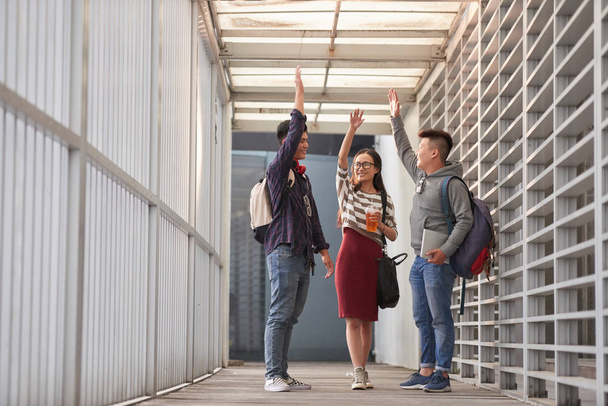 建物の廊下に木製のテラスで立ちながらハイタッチを与える陽気なアジア人留学生のグループ  - 写真・画像