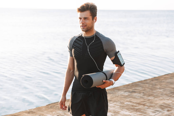 Concentrated young sportsman in earphones holding fitness mat at the beach - Фото, изображение