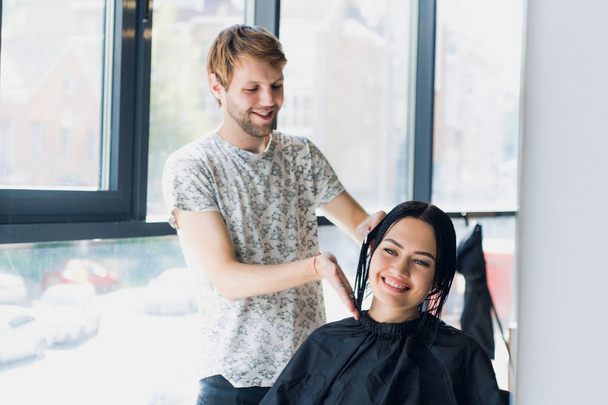 Mannelijke kapper glimlachend en praten met een klant, terwijl het maken van een nieuw kapsel aan mooie jonge brunette vrouw - Foto, afbeelding