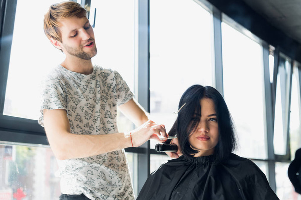 Zblízka. Ženské kadeřník narovnat hnědé vlasy žena pomocí žehlička na vlasy v salonu krásy. Ženské kadeřnice dělá účes pro mladé krásné ženy v kadeřnictví - Fotografie, Obrázek