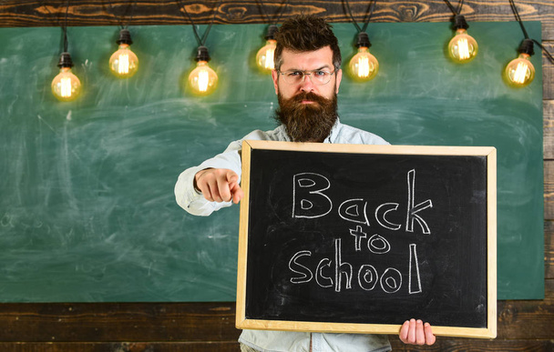 Man with beard and mustache on strict face warns students, pointing forward, chalkboard on background. Back to school concept. Teacher in eyeglasses holds blackboard with inscription back to school - Foto, imagen