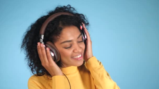 4K Smiling fashion model with headphones posing and dancing against blue background in studio. Beautiful young mixed race african female model enjoying music - Filmati, video