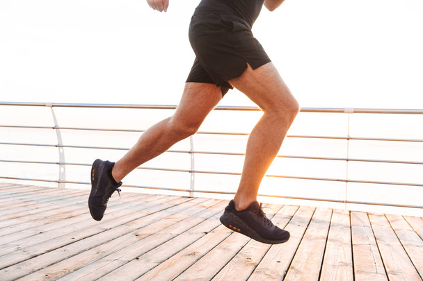 Legs closeup of active young man 20s in tracksuit running along pier or boardwalk at seaside in morning - 写真・画像