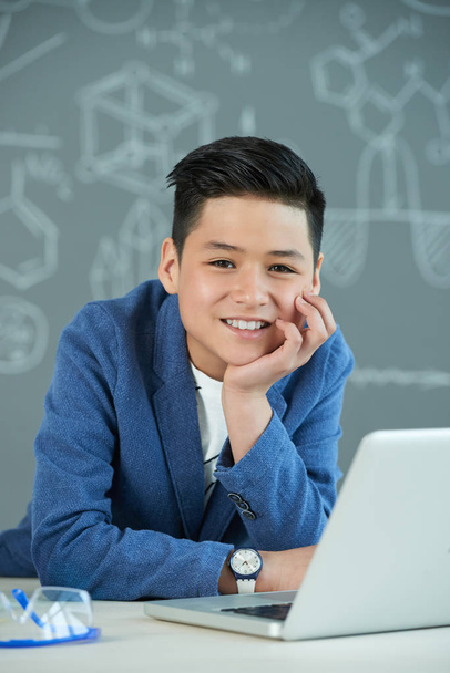 estudante asiático alegre posando para fotografia com sorriso largo enquanto descansa a cabeça na mão e sentado na mesa da escola, quadro negro com inscrições no fundo
 - Foto, Imagem
