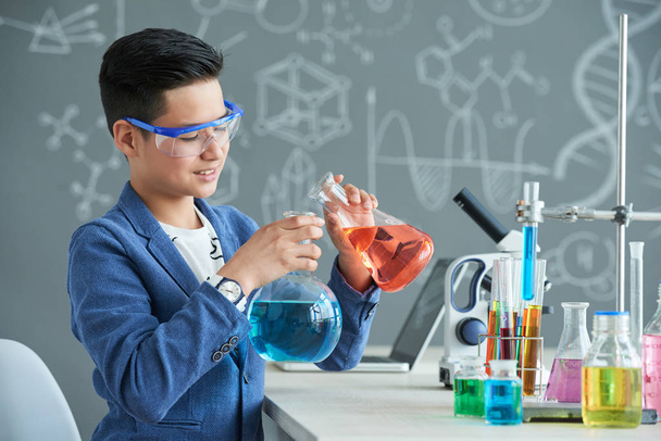 安全を身に着けている才能のある若手研究者は、化学教室で実験を遂行しながらフラスコに赤い液体を注ぐをゴーグルします。 - 写真・画像