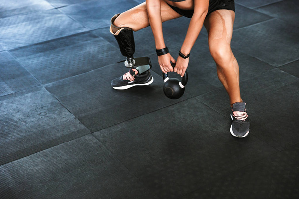 Портрет спортивной женщины-инвалида с протезом в спортивном костюме, делающей кроссфитовые упражнения и поднимающей гантель в спортзале
 - Фото, изображение
