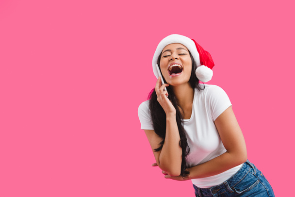 portrait of happy african american woman in santa claus hat talking on smartphone isolated on pink  - 写真・画像