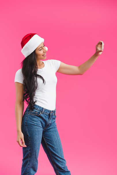 side view of smiling african american woman in santa claus hat taking selfie on smartphone isolated on pink  - Fotó, kép