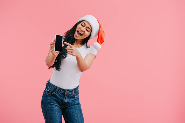 portrait de heureuse femme afro-américaine en chapeau santa claus pointant vers le smartphone avec écran vide isolé sur rose
  - Photo, image