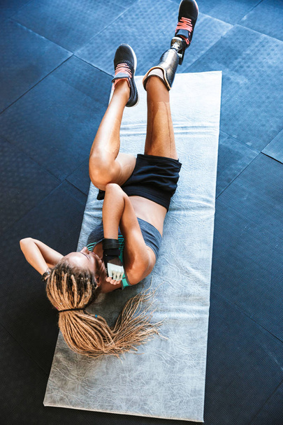 Portrait from above of fitness disabled sportsgirl with prosthesis in tracksuit lying on mat and doing criss cross crunches in gym - Фото, изображение