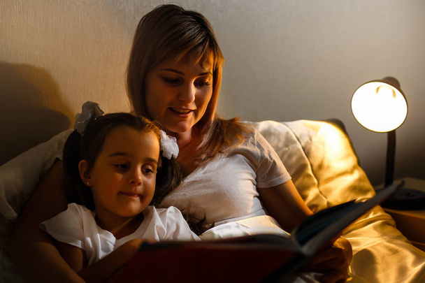 молодая мать и маленькая девочка читают книгу в постели перед сном
 - Фото, изображение